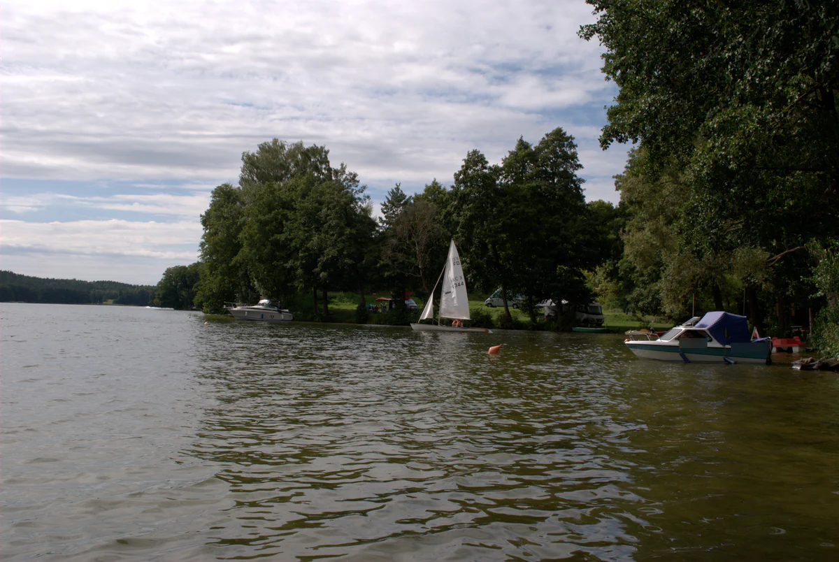 Panorama des Ukiel-Sees vom Wasser aus: eine Jolle unter Segeln, vertäute Motorboote und das bewaldete Campingufer.